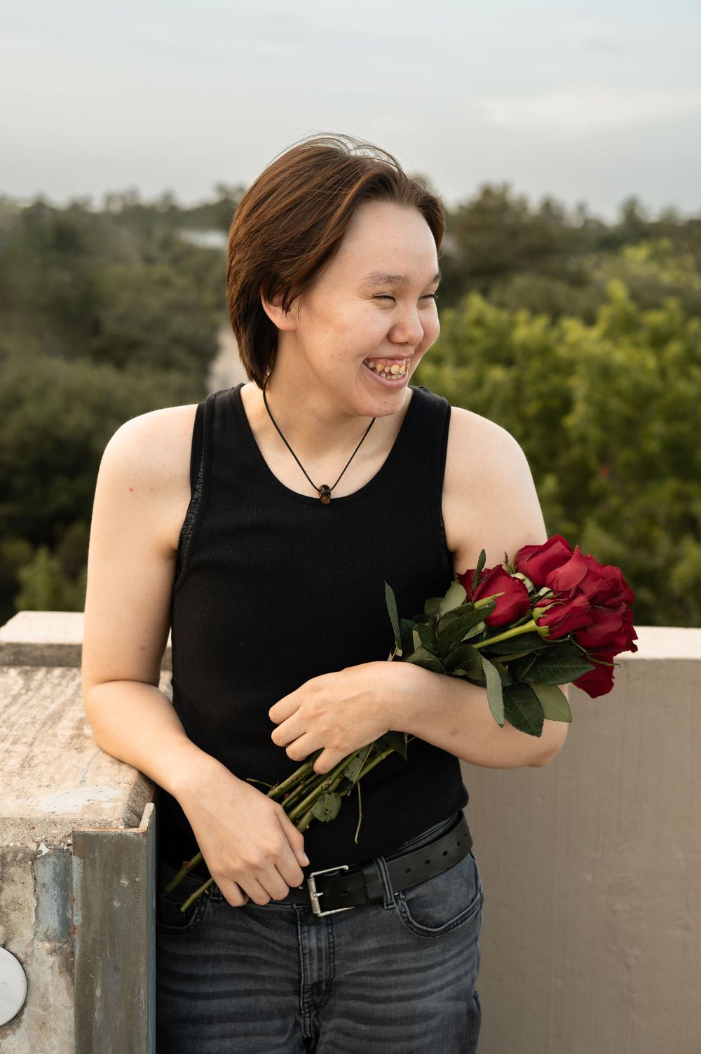 Someone in a black tank top holds red roses while smiling on a rooftop with greenery in the background.