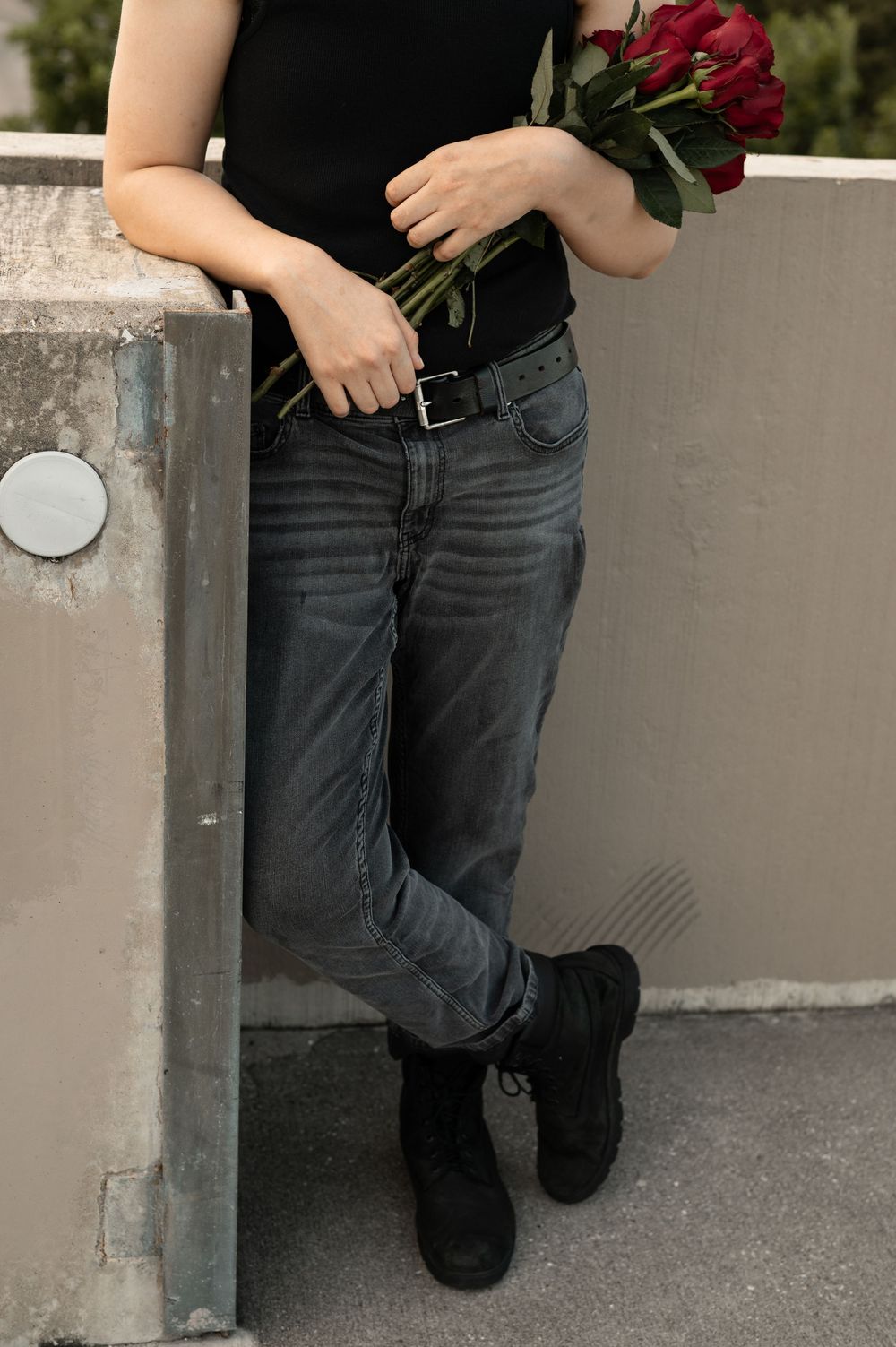 Close-up view of person wearing black jeans and boots holding red roses against a concrete wall.