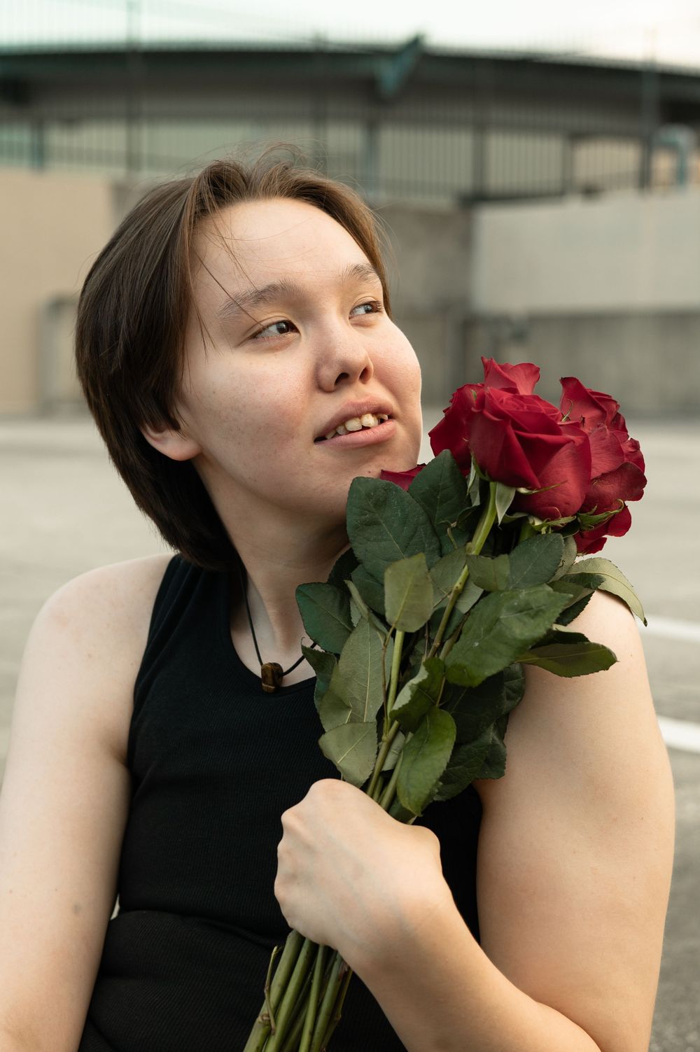 Someone in a black sleeveless top holds vibrant red roses while smiling against an urban background.
