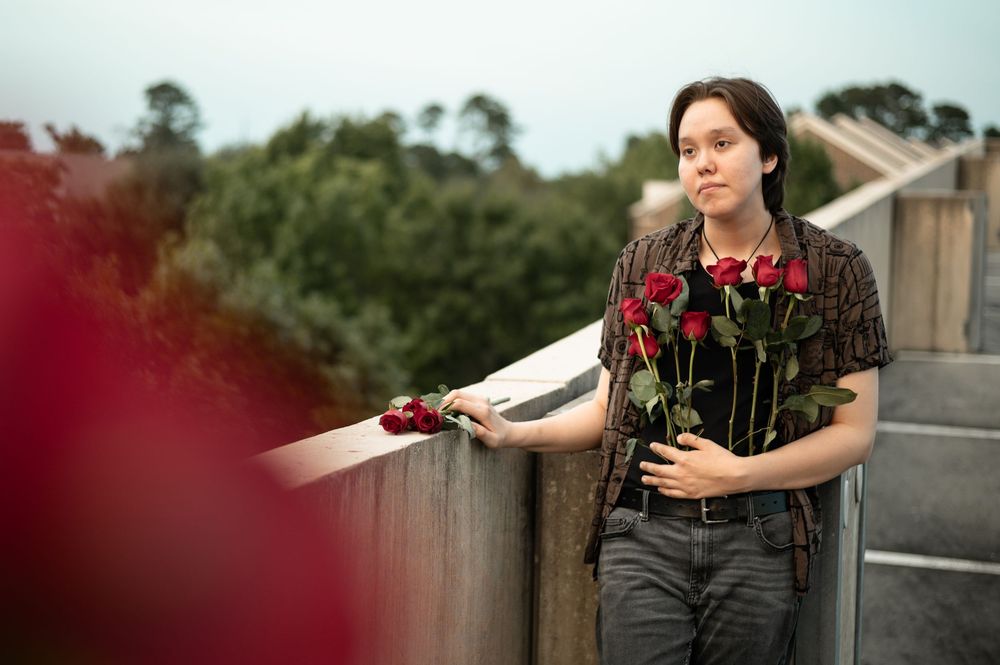 Someone holding red roses stands by wooden railing on bridge in outdoor setting with trees in background.