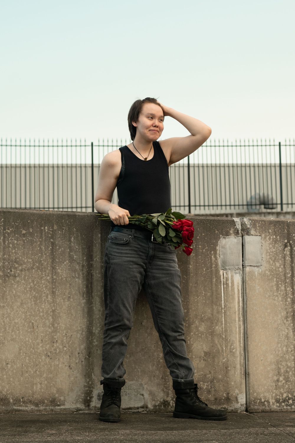 A figure in a black tank top and dark jeans leans against a concrete wall holding red roses.