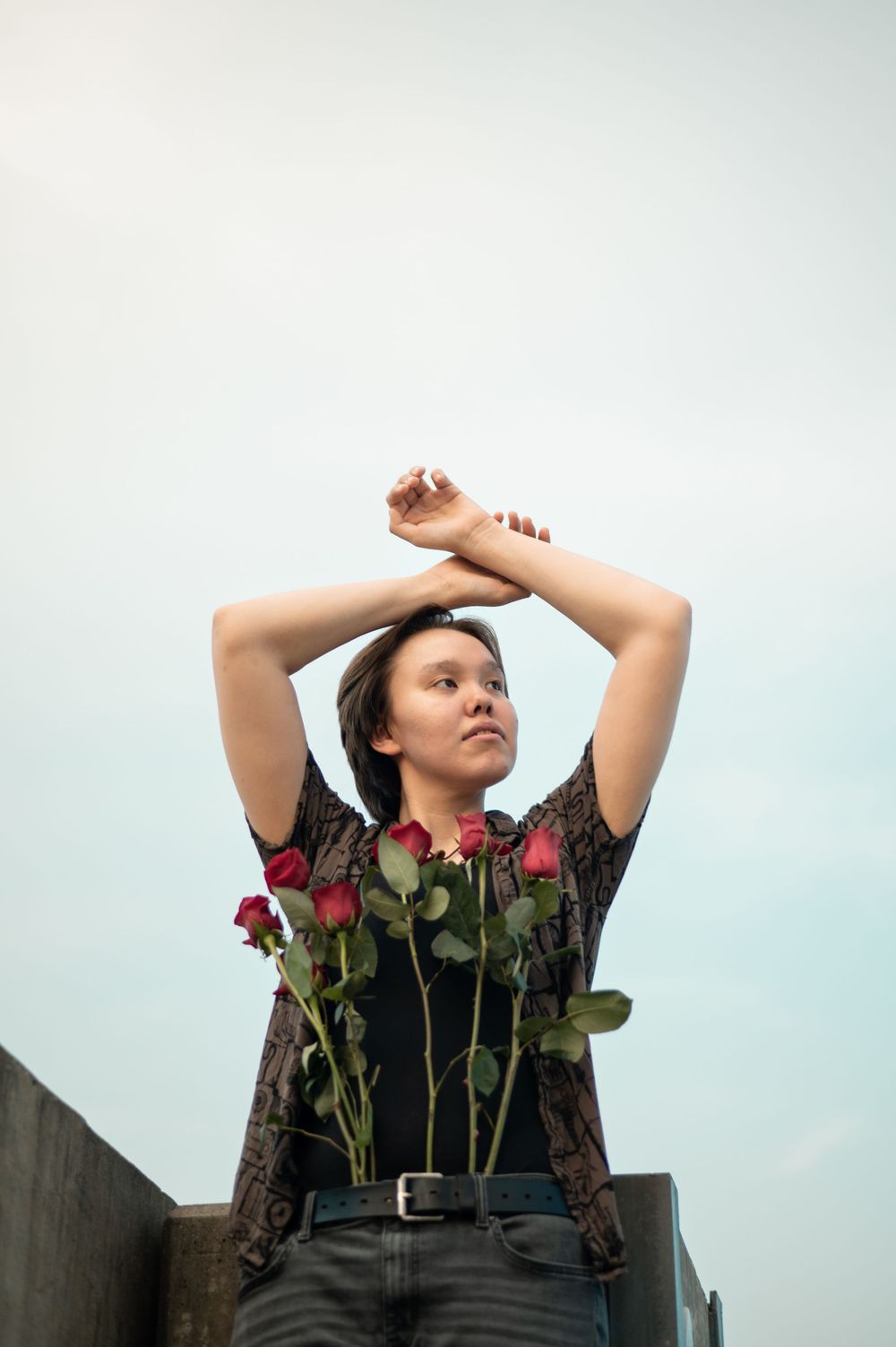 A person in a dark top poses against a light sky with red roses, lifting their arms above their head in an artistic stance.