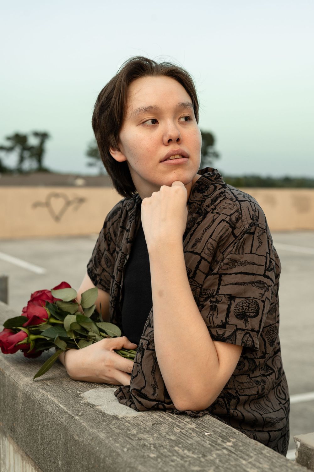 Someone in a brown patterned dress lies on a concrete barrier with a rose, creating a moody outdoor portrait series.