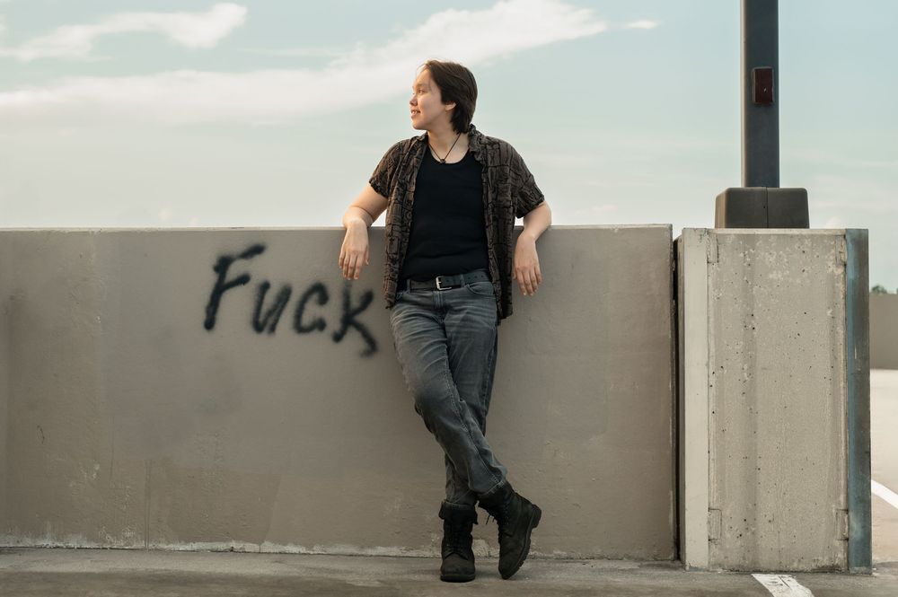 Person in black attire posing against a light concrete wall in an urban setting.