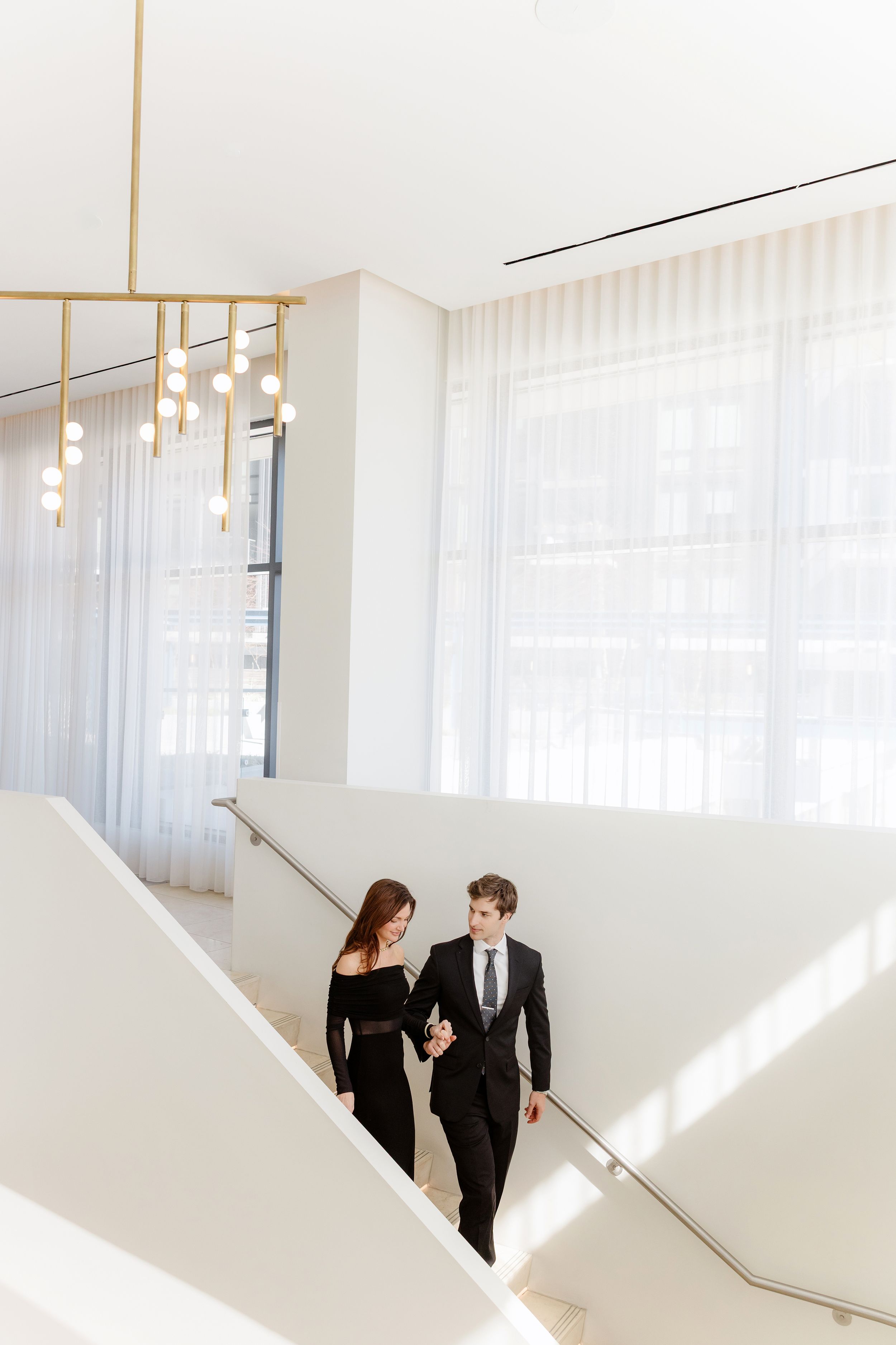 A couple in formal attire walking up a modern white staircase with sheer curtains and pendant lights in a bright interior.