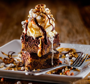 Decadent chocolate brownie topped with whipped cream, caramel sauce and chopped nuts on a white plate.