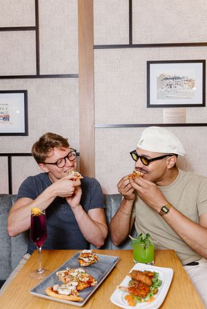 Two friends share a meal at a restaurant with sandwiches and drinks while sitting at a wooden table with framed art on walls.