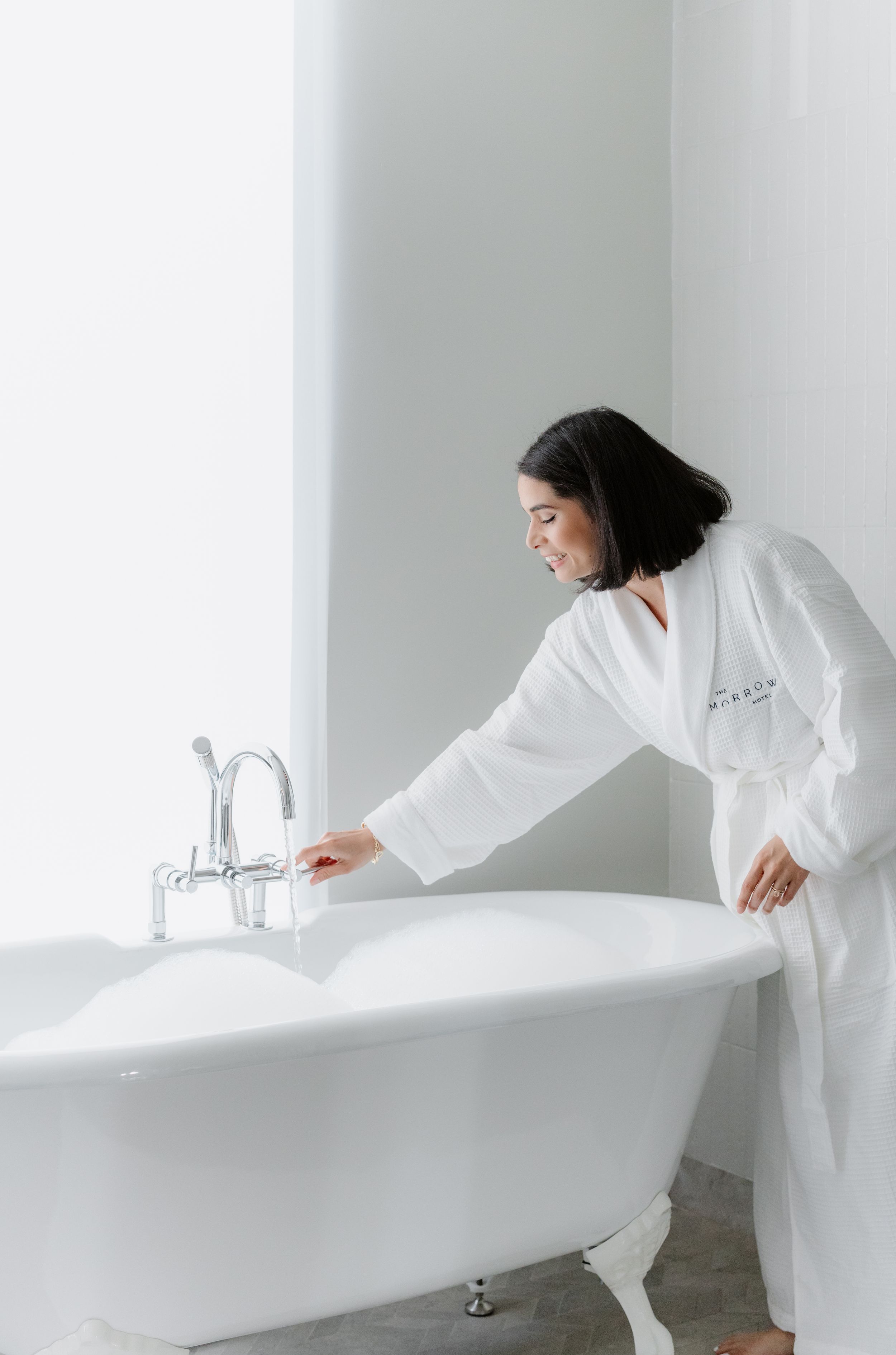 Someone in a white bathrobe checks the water temperature in a classic freestanding white bathtub in a minimalist bathroom.