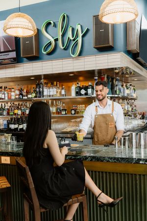 Restaurant bar interior with neon 'Sly' sign featuring modern pendant lights and bartender serving customer at marble counter.