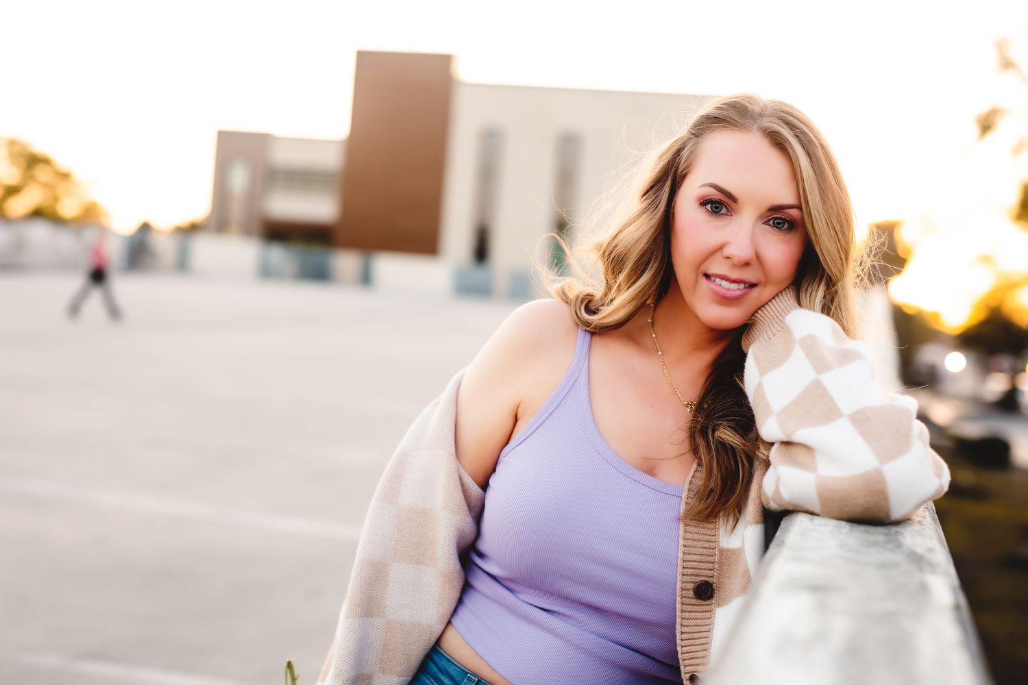 Woman wearing purple tank top and beige cardigan smiles brightly outdoors at sunset.