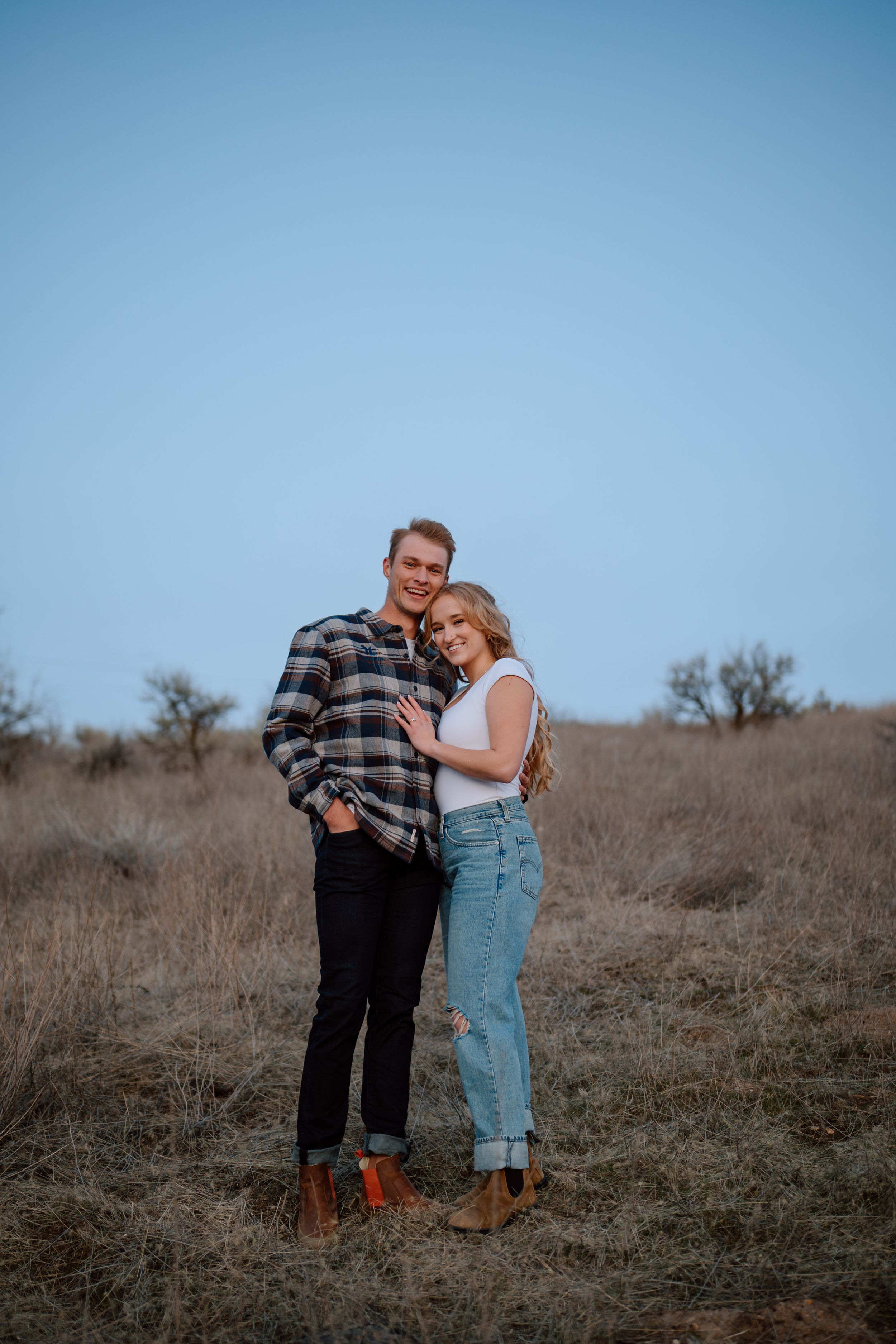 A couple in casual attire embracing on a hillside during golden hour against a blue sky backdrop.