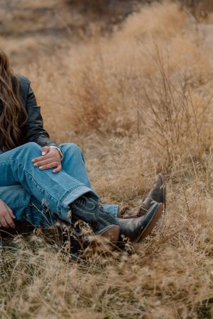 Person wearing denim jeans and black boots sits in golden dry grass during autumn season.