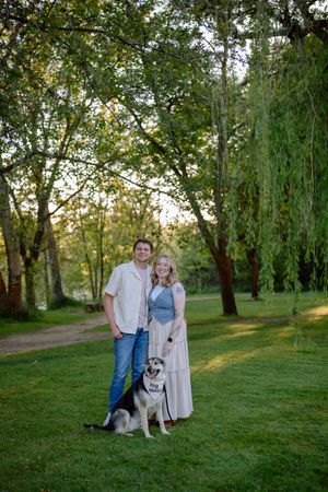 A couple stands with their husky dog on a grassy lawn surrounded by willow trees at sunset.