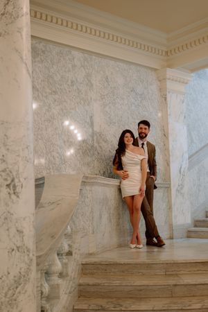 A couple embraces on an elegant marble staircase in a bright white interior with ornate crown molding details.