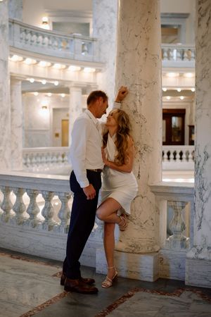 Romantic couple poses intimately by marble columns in an elegant historic building interior with ornate balustrades.