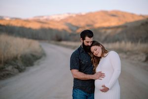 A couple embraces on a dirt road with golden mountains and rolling hills in the background at sunset.