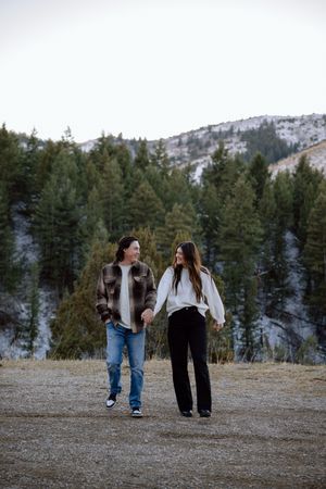 A couple takes a romantic stroll in the outdoors with snow-capped mountains and evergreen trees in the background.