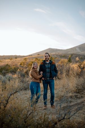 A couple embraces while standing in a desert landscape with brown brush and mountains in the background during golden hour.