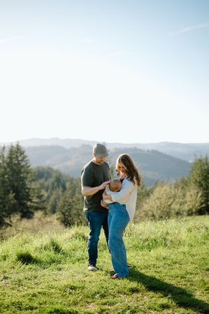A couple stands together on a grassy hilltop overlooking mountain ranges and evergreen forests on a sunny day.