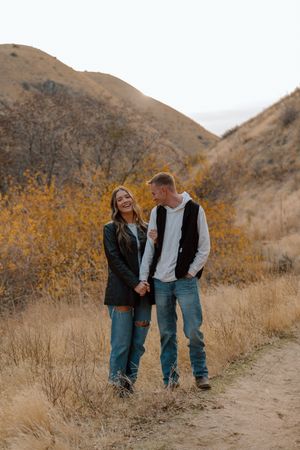 A couple wearing casual attire walks together on a dirt path surrounded by autumn-colored hills and dry grass.