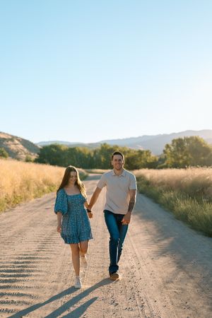 A couple walks together on a dirt road during golden hour with mountains and tall grass in the background.