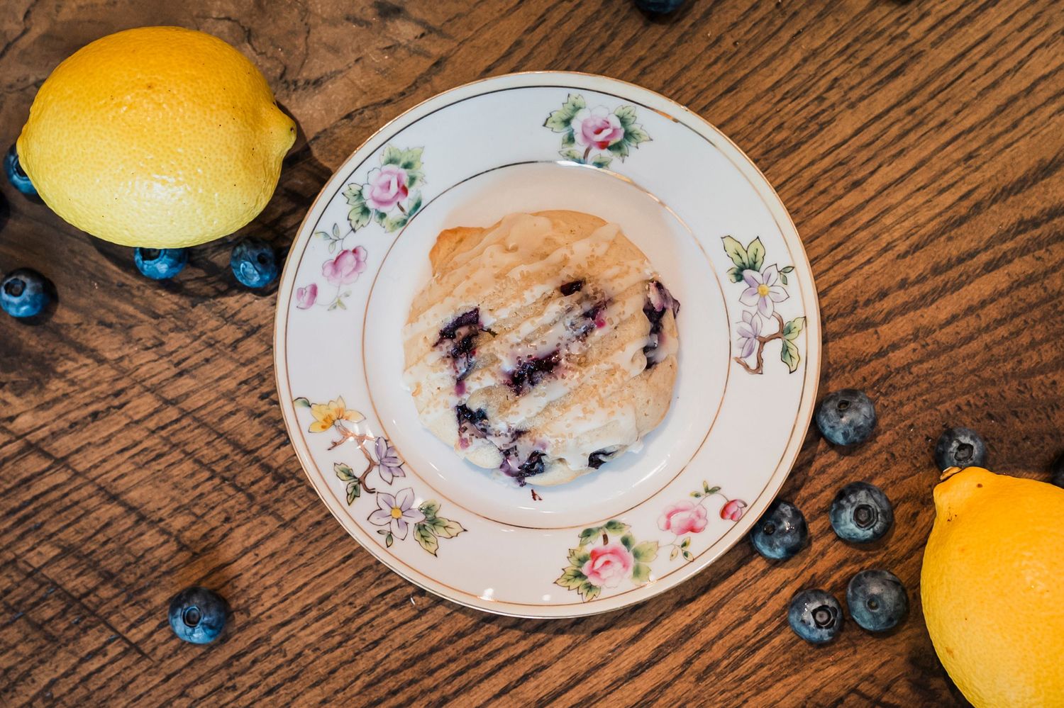 A blueberry muffin on a floral plate with fresh blueberries and oranges scattered on wooden table.