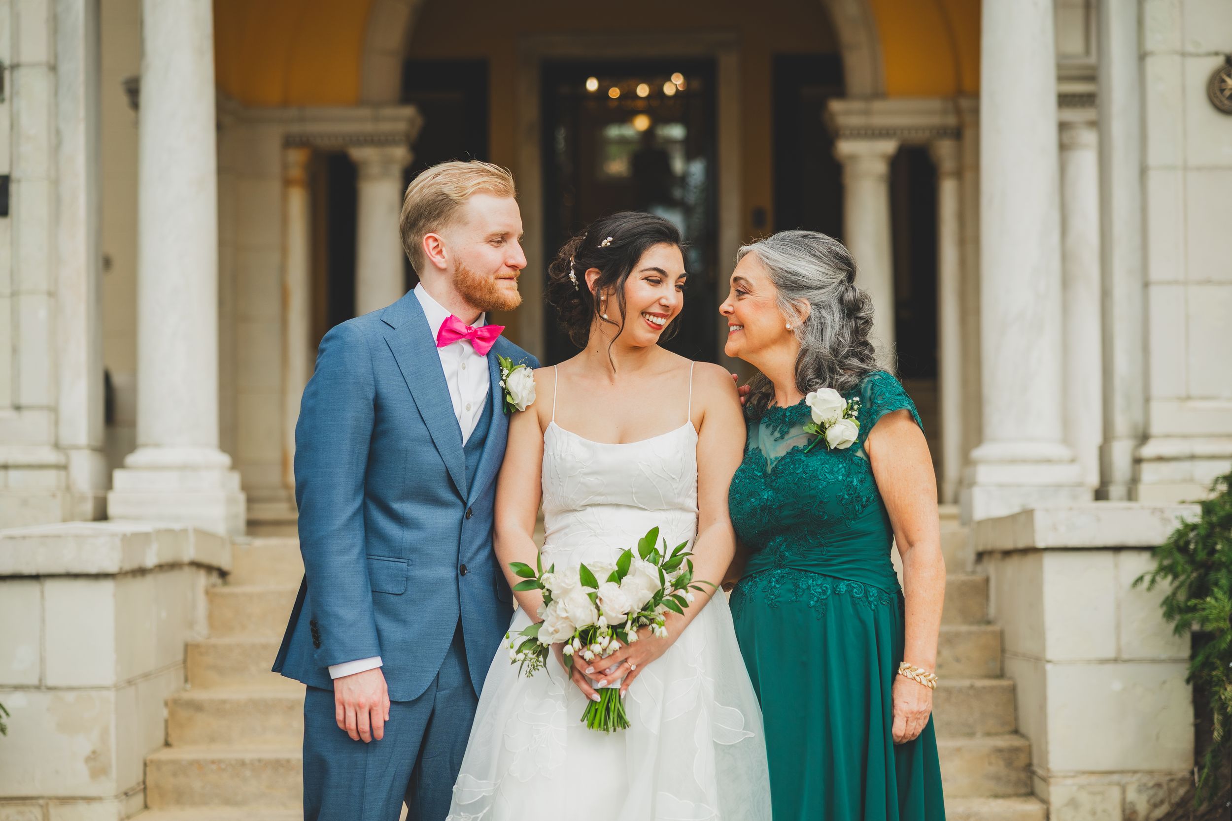 Wedding party members stand together in emerald green dresses and blue suits while holding white floral bouquets.