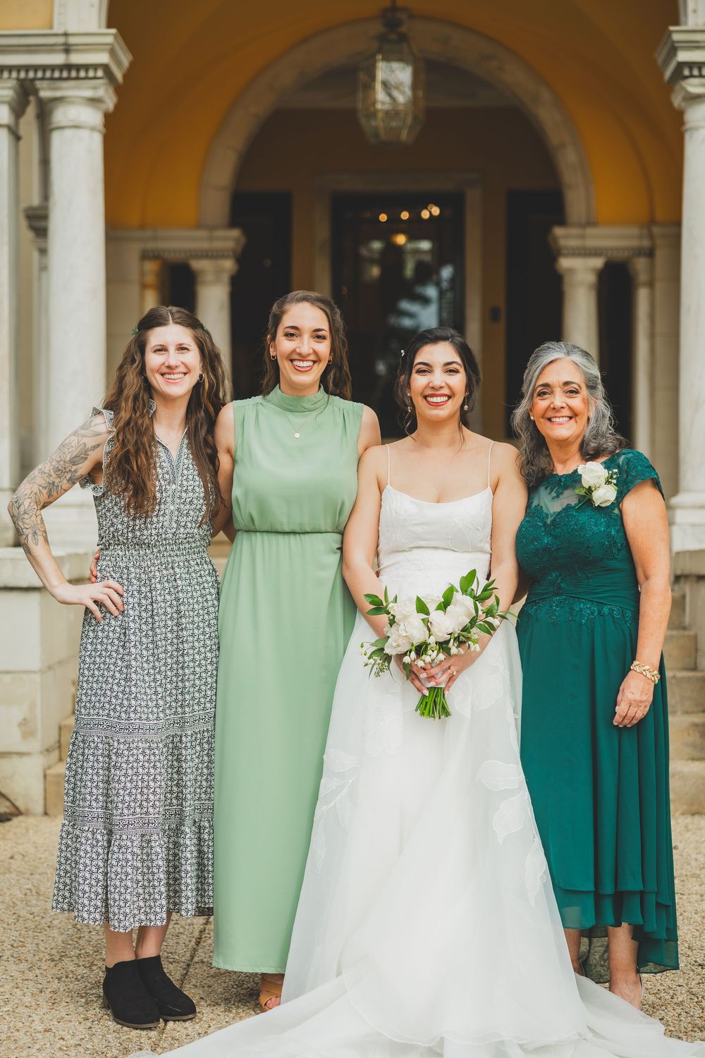 Wedding party wearing dresses in shades of sage green, emerald, and patterned grey pose together under an arched entrance.