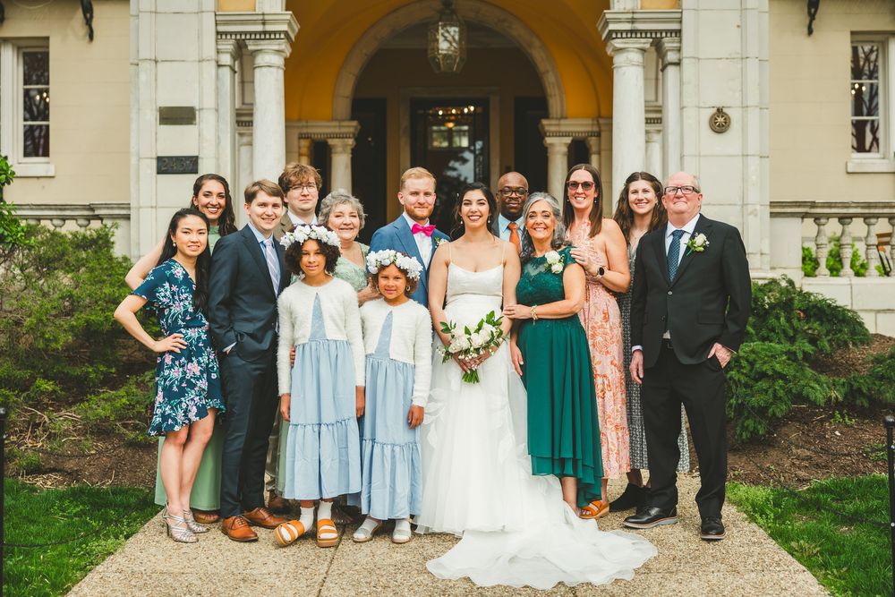 A wedding party poses together on the steps of an elegant stone building with columns and arched entrance.