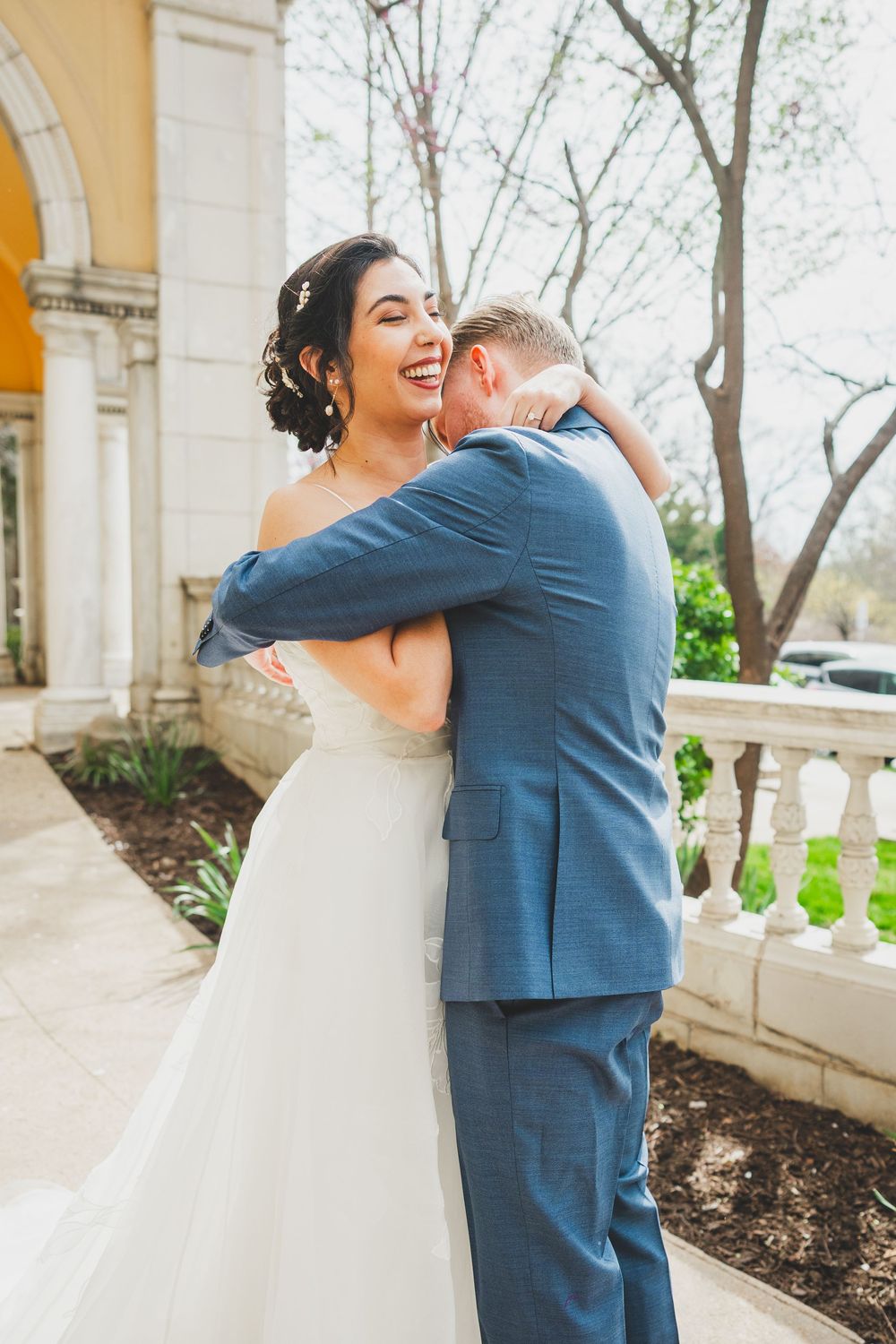 A happy couple in wedding attire embrace and laugh together on the grounds of an elegant stone building at the Josephine Butler Parks Center