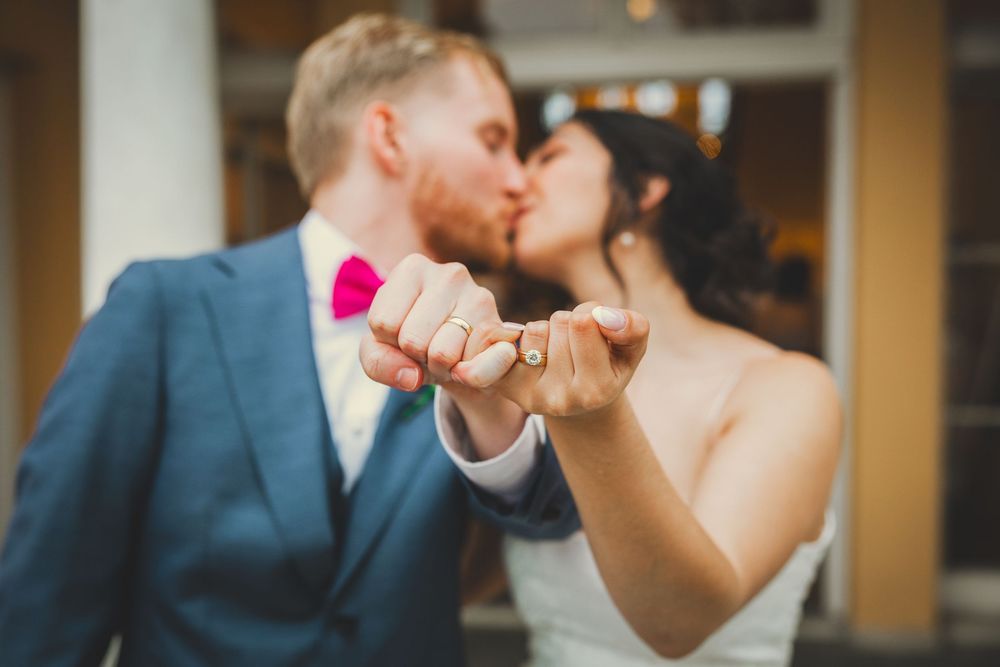 A bride and groom share a romantic kiss while holding up their hands displaying wedding rings.
