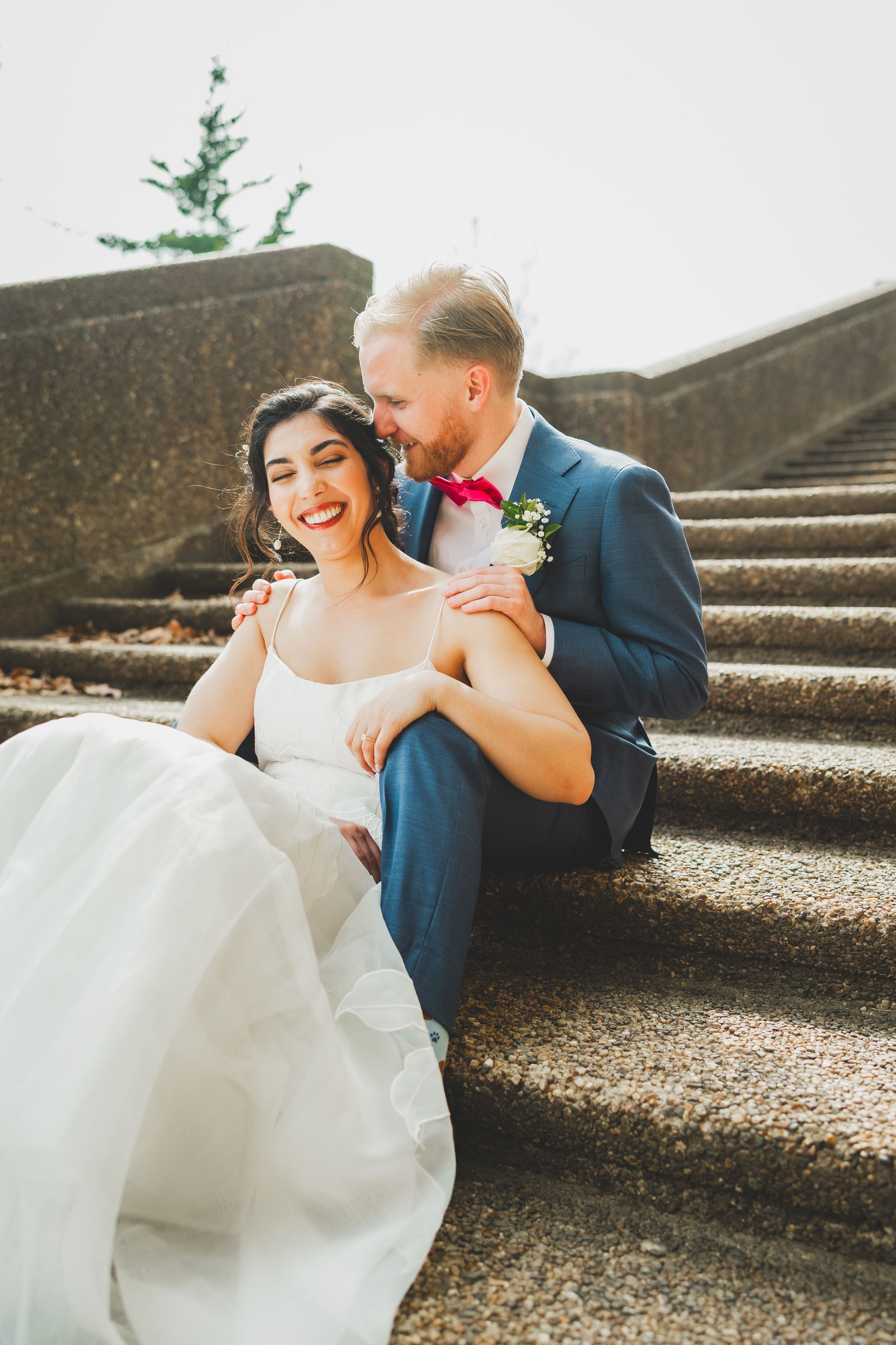 A joyful wedding couple sharing a tender moment while sitting on concrete steps in warm sunlight.