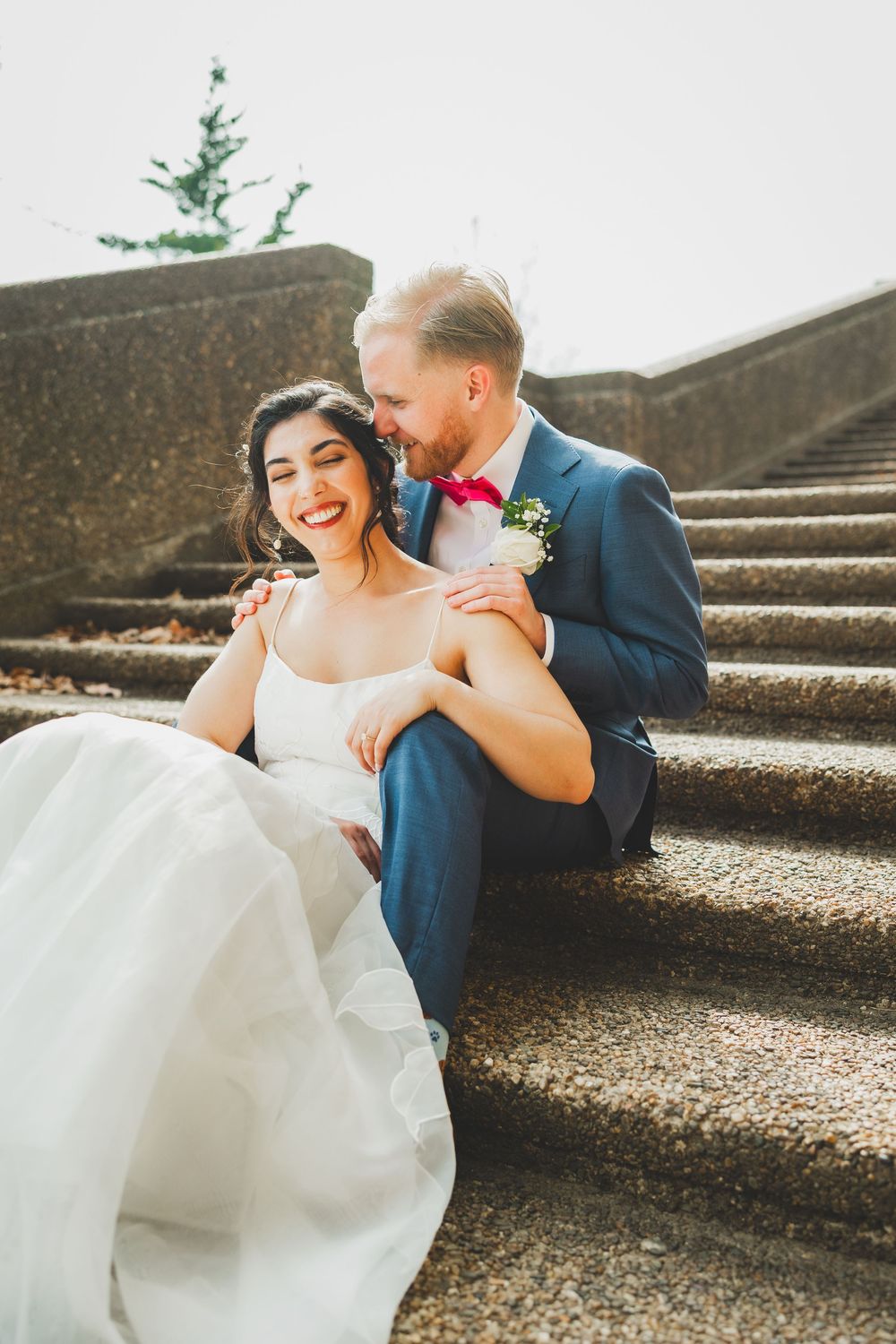 A joyful wedding couple sharing a tender moment while sitting on concrete steps in warm sunlight.