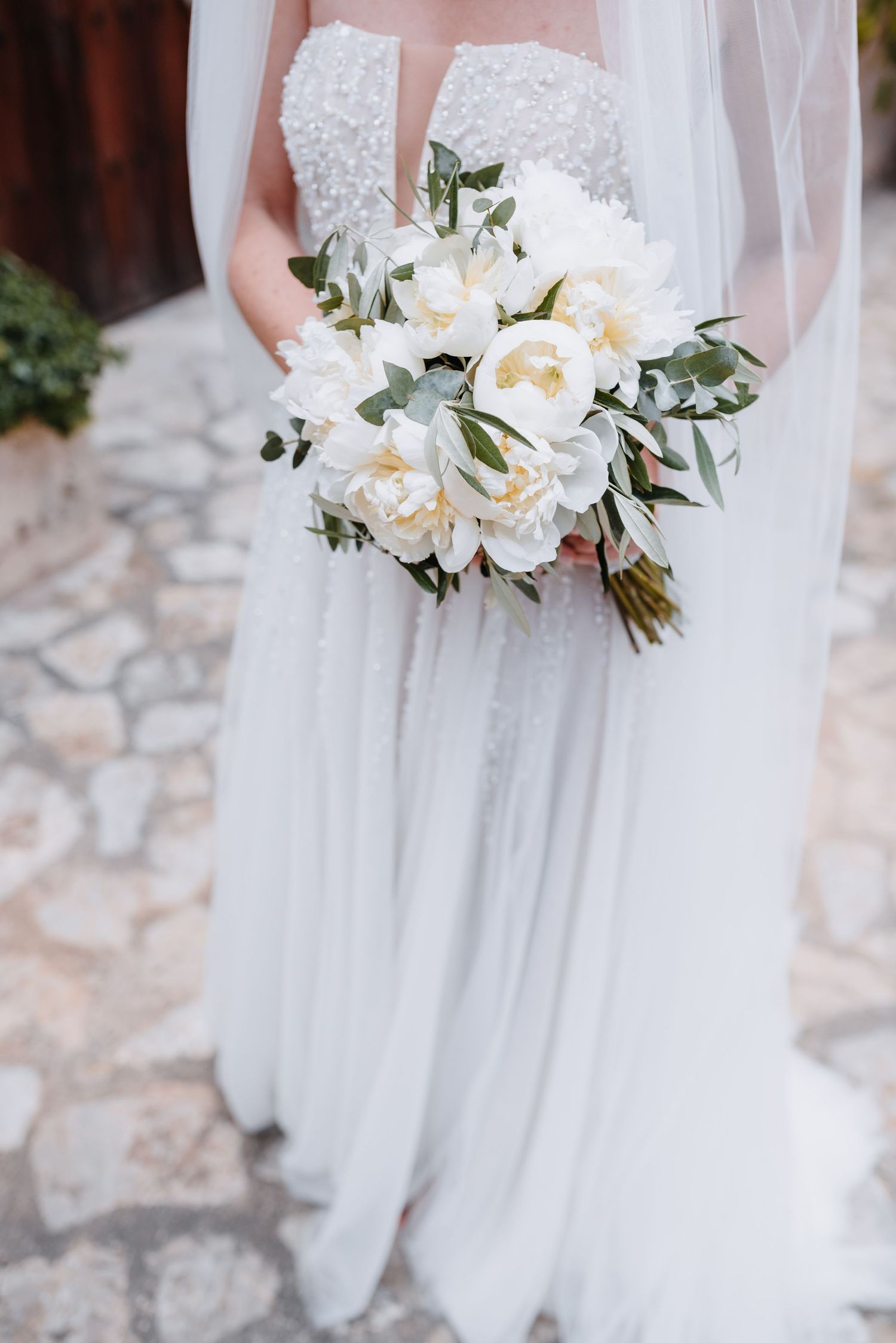 Detailbild eines zarten weißen Brautstrauß' mit Pfingstrosen und Eukalyptusblättern, gehalten vor einem fließenden weißen Brautkleid.