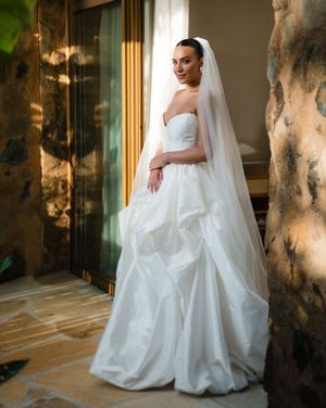 A bride in a flowing white gown and veil poses elegantly against a wall with dramatic window lighting.