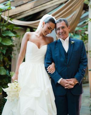 A bride in a white strapless wedding gown stands with a companion in a navy suit at an outdoor garden venue.
