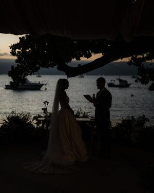 Silhouettes of a romantic wedding couple exchange vows under a tree at sunset with boats anchored in the background.