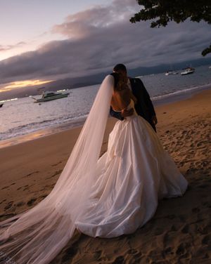 A couple in wedding attire embraces on a sandy beach at sunset with dramatic cloudy skies over the ocean.