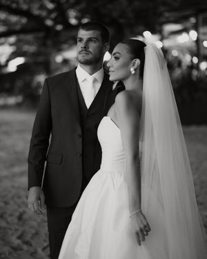 Classic black and white wedding portrait showing a bride in simple gown with veil and groom in suit standing outdoors.