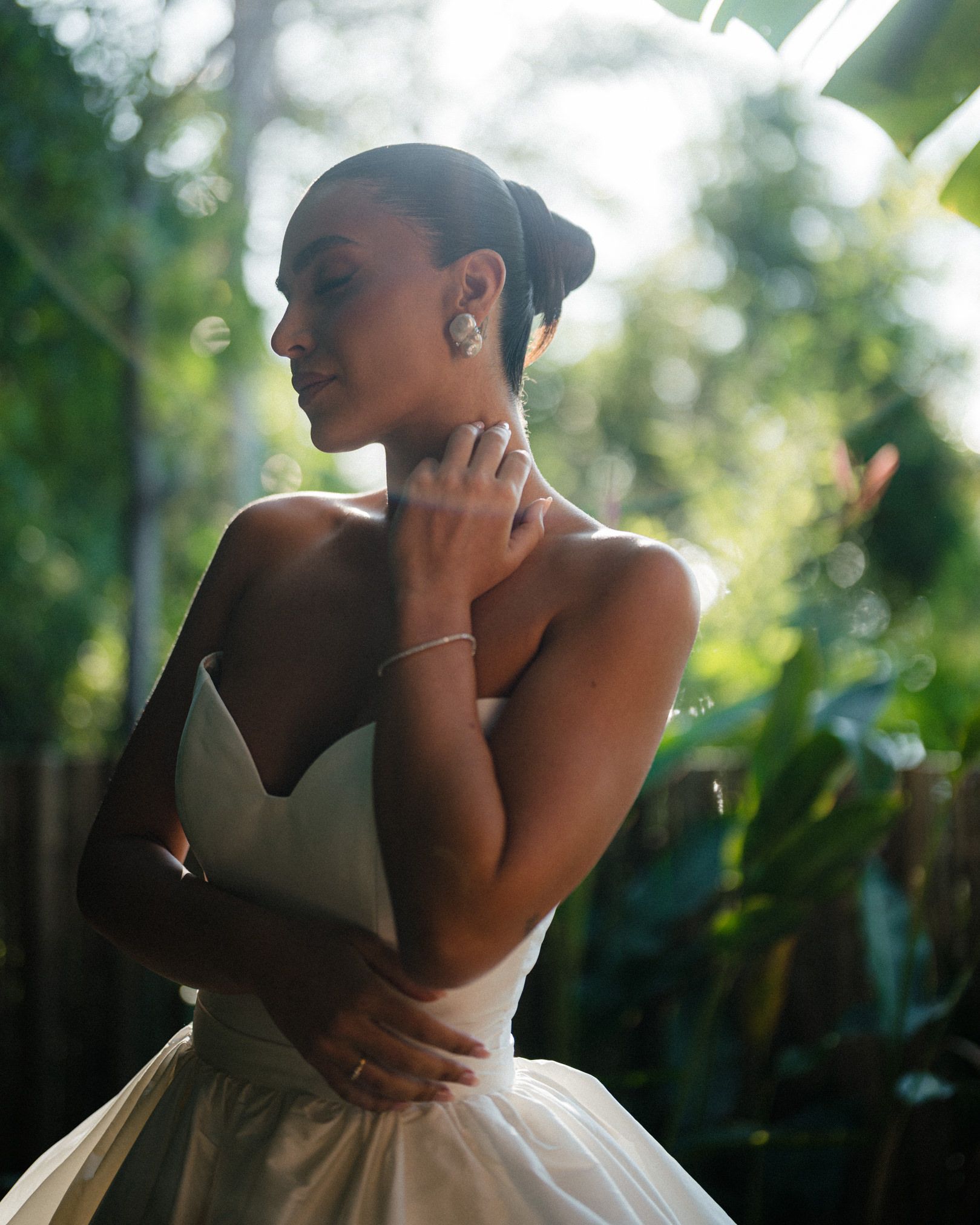 A silhouette of an elegant figure in a white wedding dress stands in soft sunlight among green foliage.