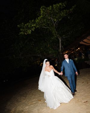 A newlywed couple walks along a dimly lit beach path under trees with twinkling lights in the evening.