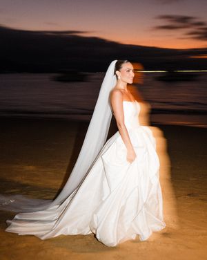 A bride in a white wedding dress sits on a sandy beach during sunset with a flowing veil and dramatic lighting.