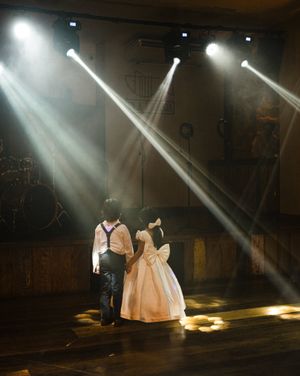 Dramatic spotlights illuminate a couple in formal attire standing on a dark wooden stage during what appears to be a performance.