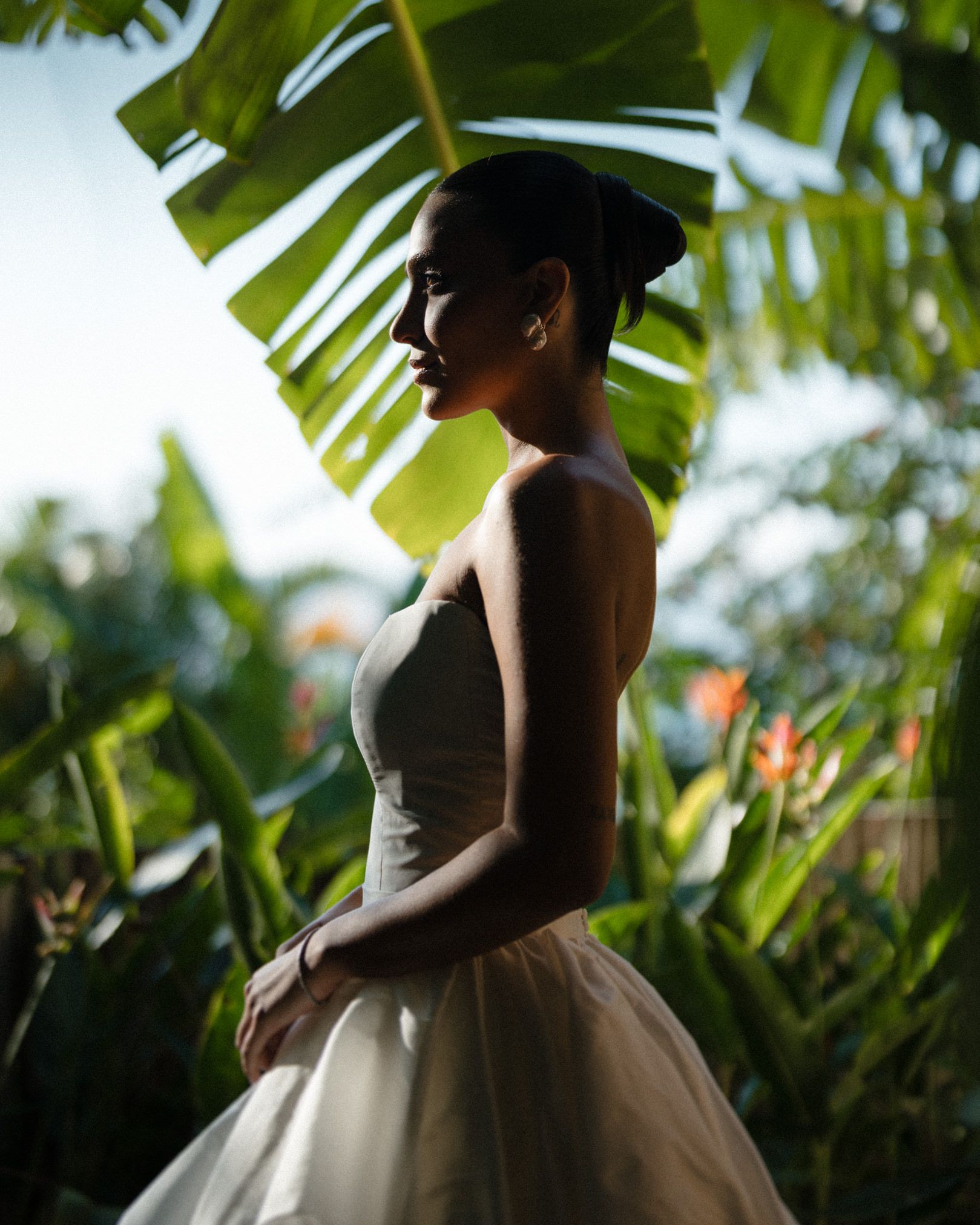 A silhouetted figure in an elegant ballgown stands beneath tropical palm fronds in a romantic garden setting.