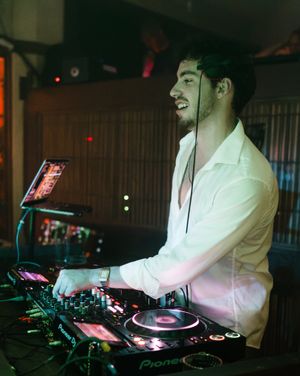 A DJ performs at a club setting with vinyl turntables and mixing equipment under atmospheric lighting.
