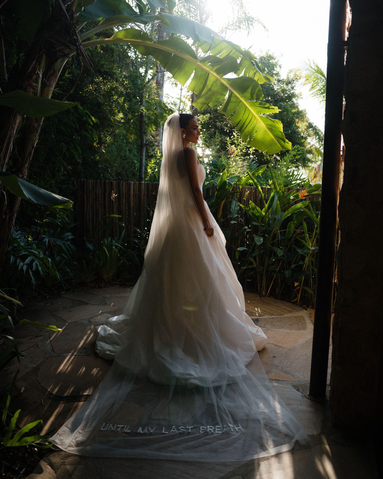 Beautiful white wedding dress with flowing train captured in motion against lush tropical garden backdrop in natural light.