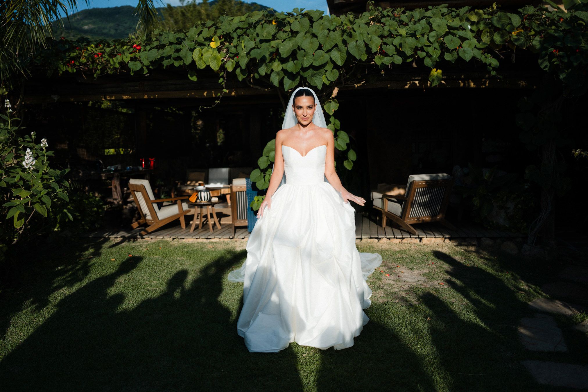 A bride in a flowing white wedding dress walks through a sun-dappled garden pathway surrounded by lush greenery.