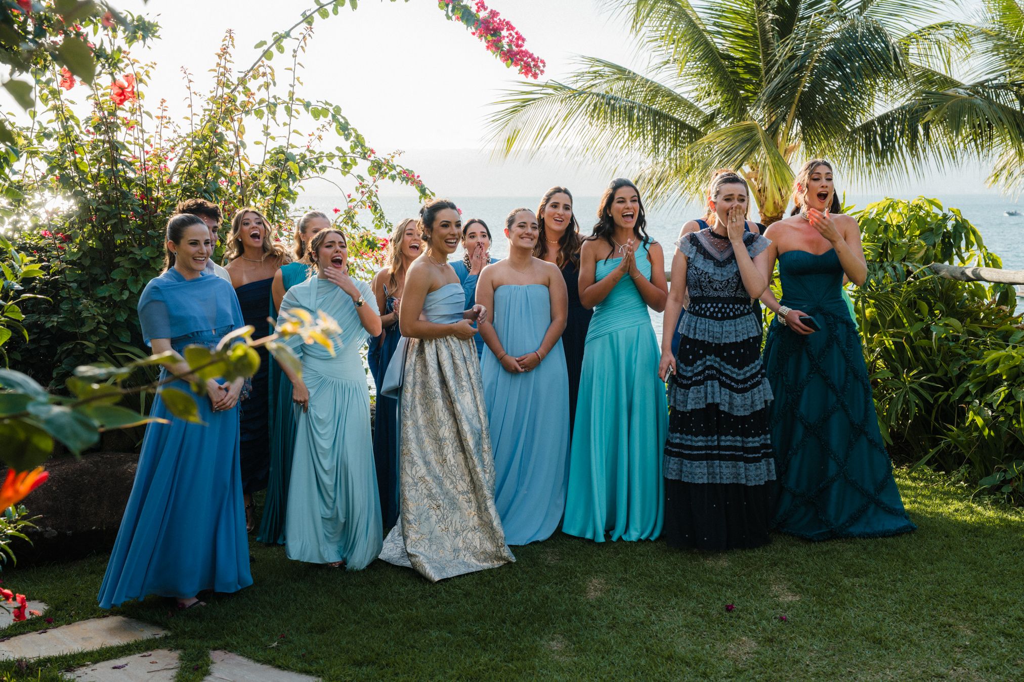 Wedding party members in coordinated blue and turquoise dresses pose together on a tropical lawn with palm trees.