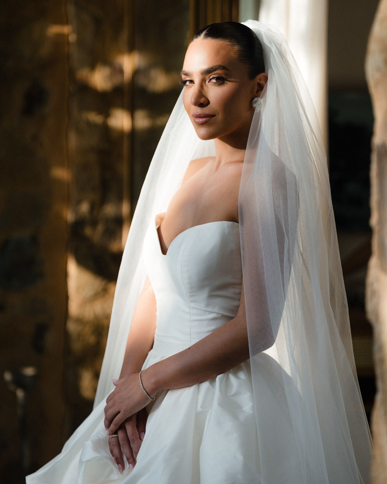 A bride in a classic white strapless wedding gown poses elegantly with a flowing veil in a softly lit indoor setting.