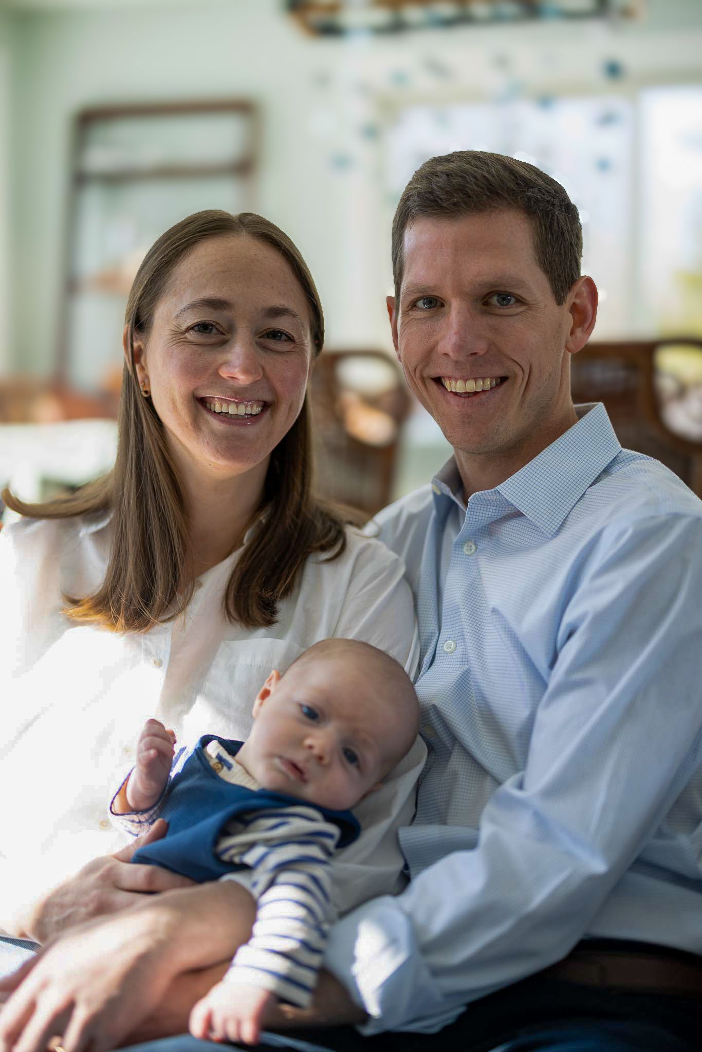A couple smiles while holding their sleeping baby in a blue outfit.